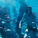 Lanky blue cat-people swim underwater next to a manta ray-looking thing.